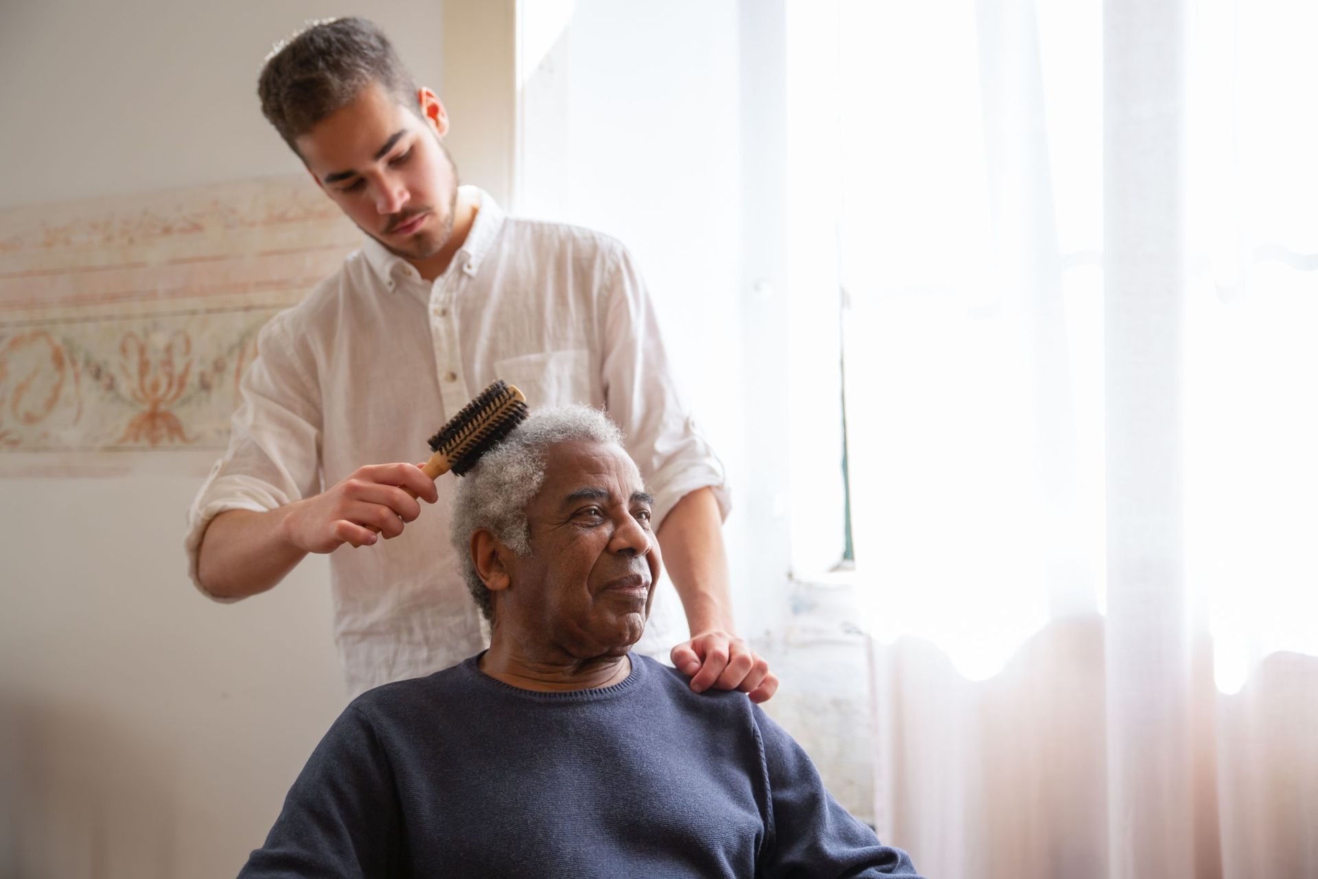 A carer brushing an older man's hair by the window
