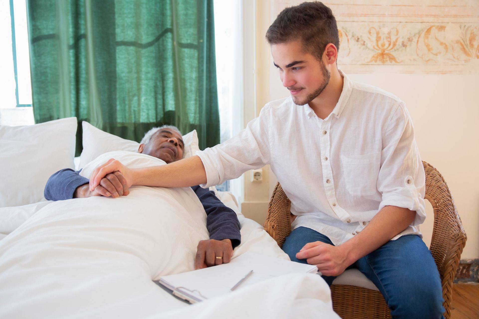 A carer sitting at the bedside, resting a hand on an older man in bed