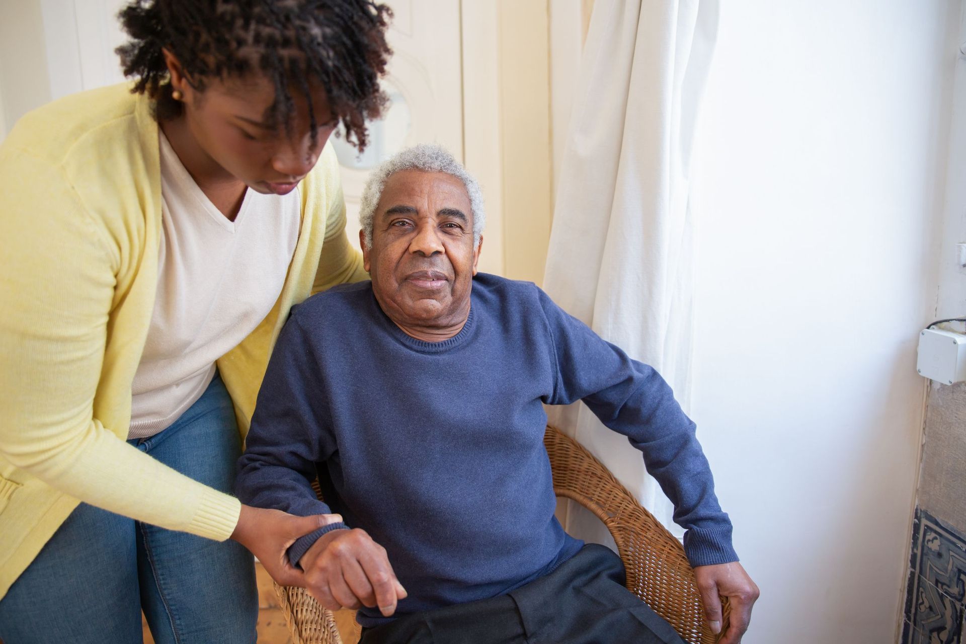 A carer in a yellow cardigan helping an older man in a navy jumper settle into a wicker armchair