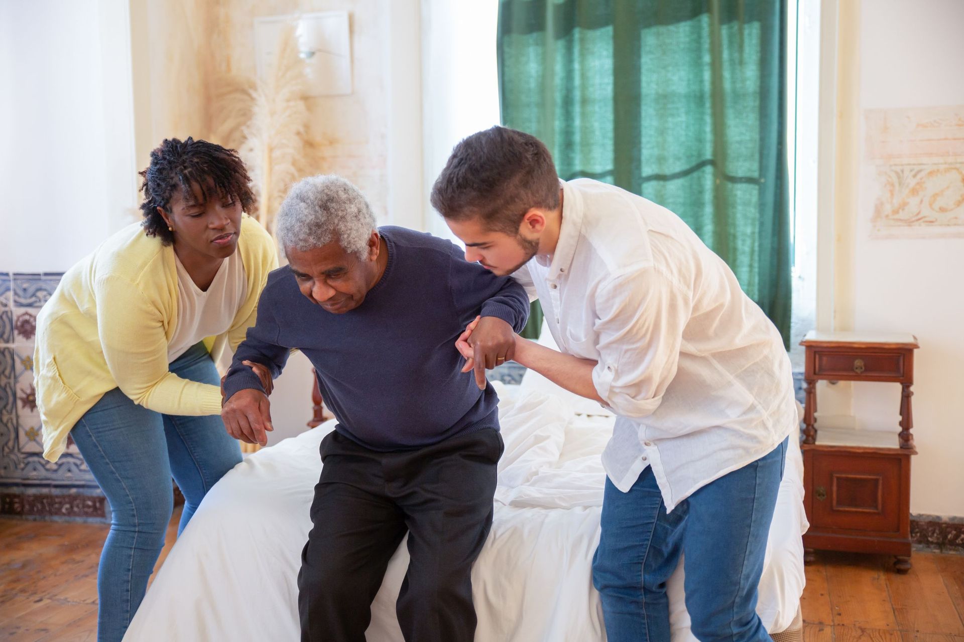 Two carers helping an older man stand up from the edge of his bed