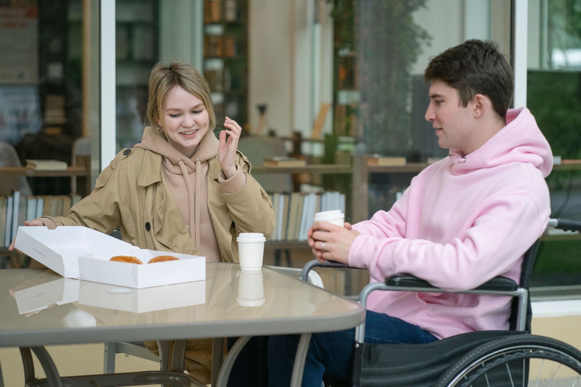 Two young people chatting over coffee at a café table, one using a wheelchair