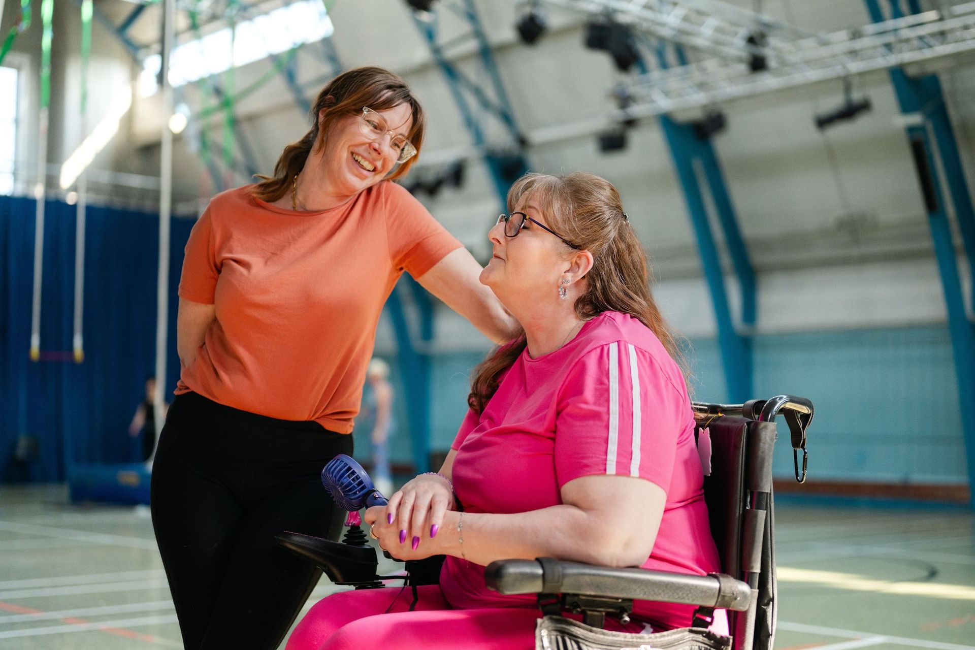 Two women laughing together in a sports hall, one using a wheelchair